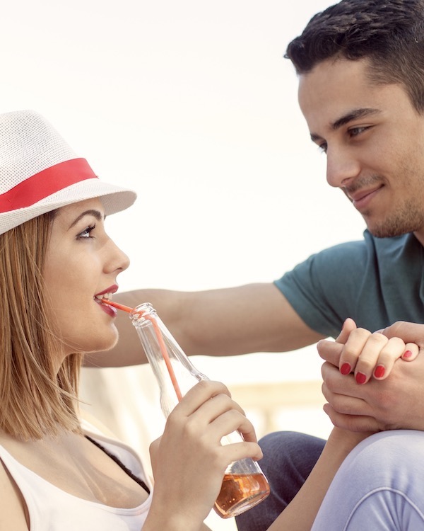 Ein junges Paar flirtet am Strand durch Blickkontakt, Händchen halten und schüchternem Lächeln. 