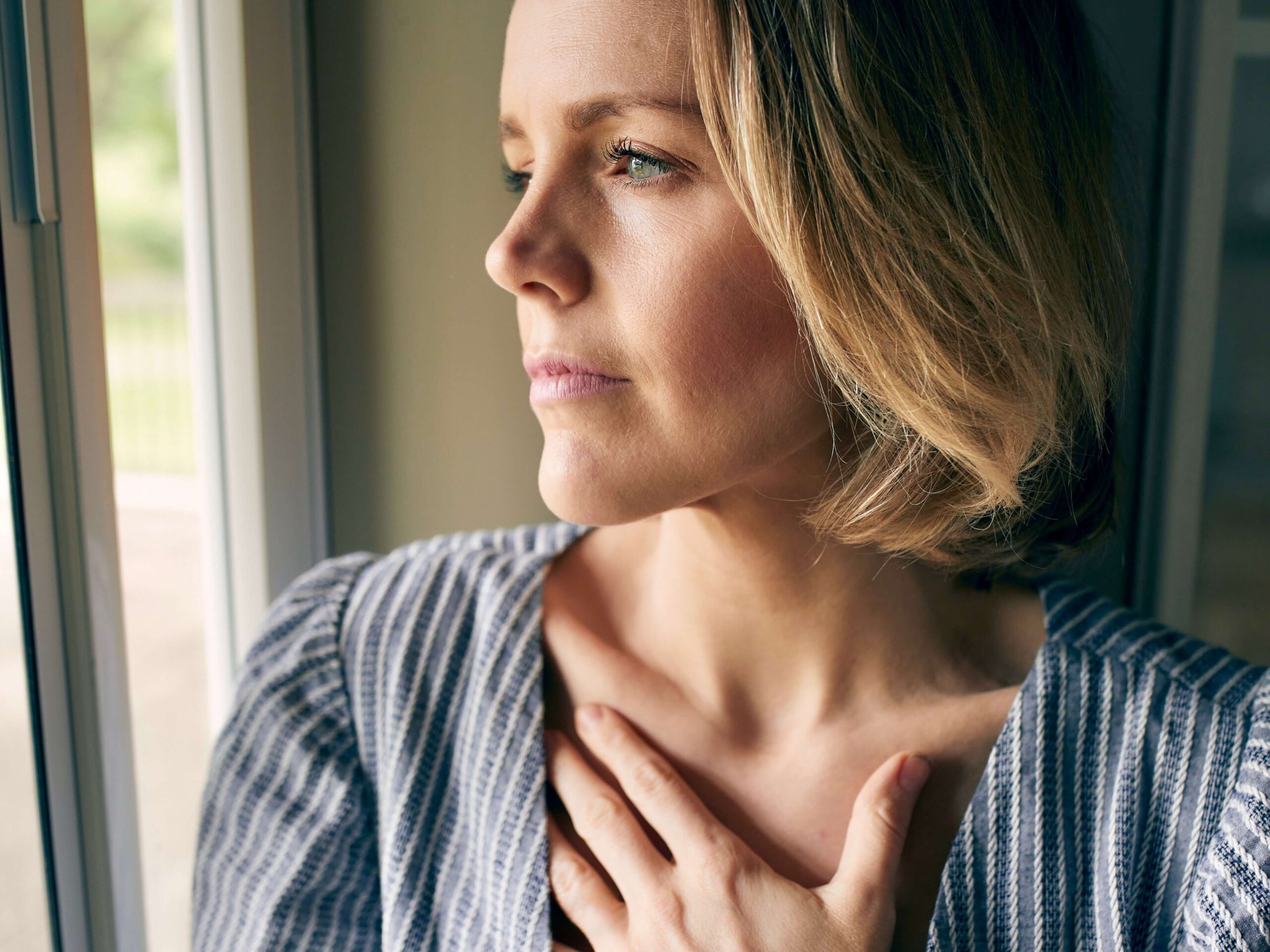 Eine nachdenkliche Frau blickt aus dem Fenster, ihre Hand liegt auf dem Herz und in ihrem Kopf kreist die Frage: Warum kann ich nicht einfach loslassen?